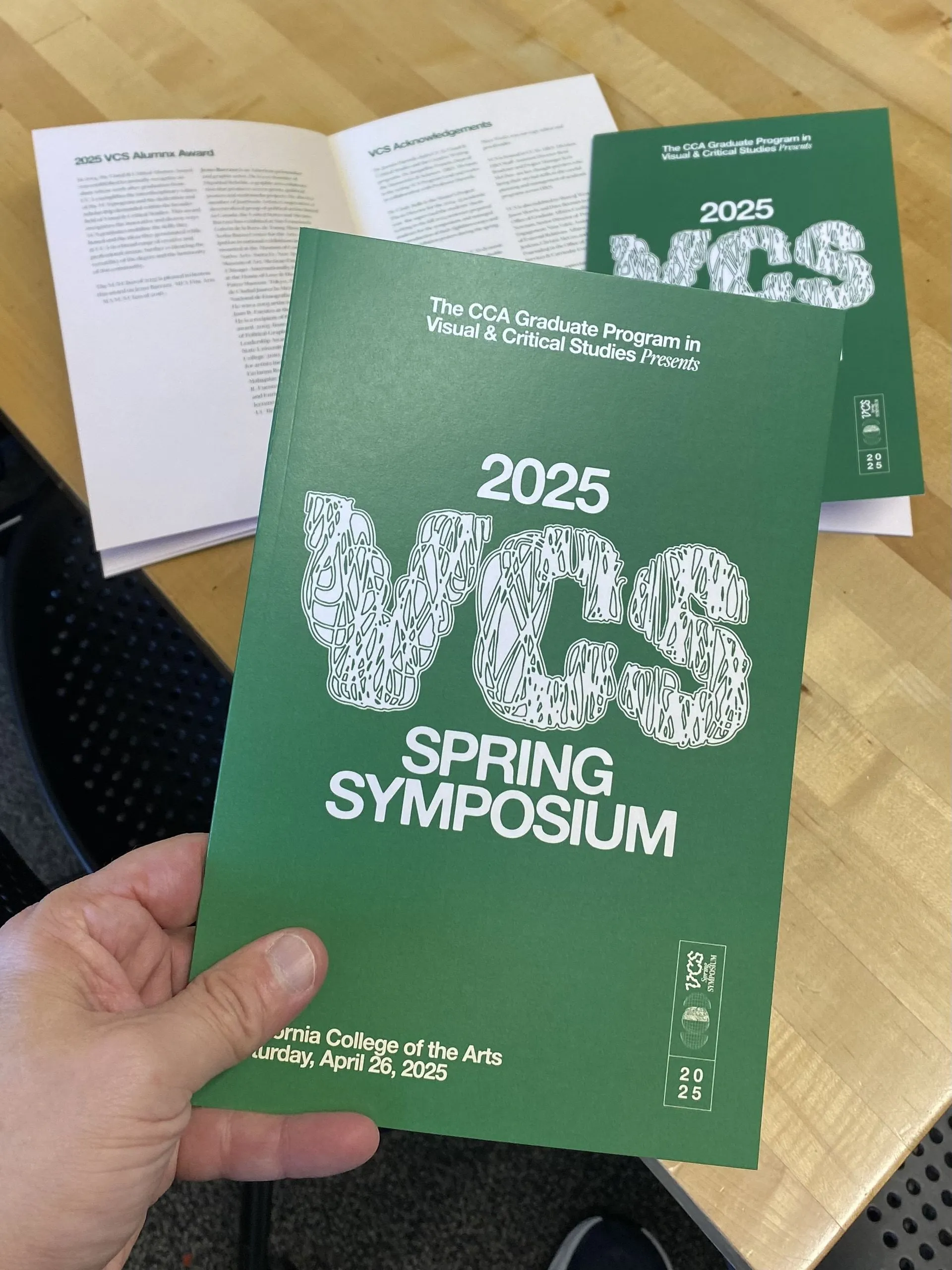 A hand holds a dark green VCS Spring Symposium program booklet printed with large white textured lettering; a second open copy and another booklet rest on a wood table behind it.