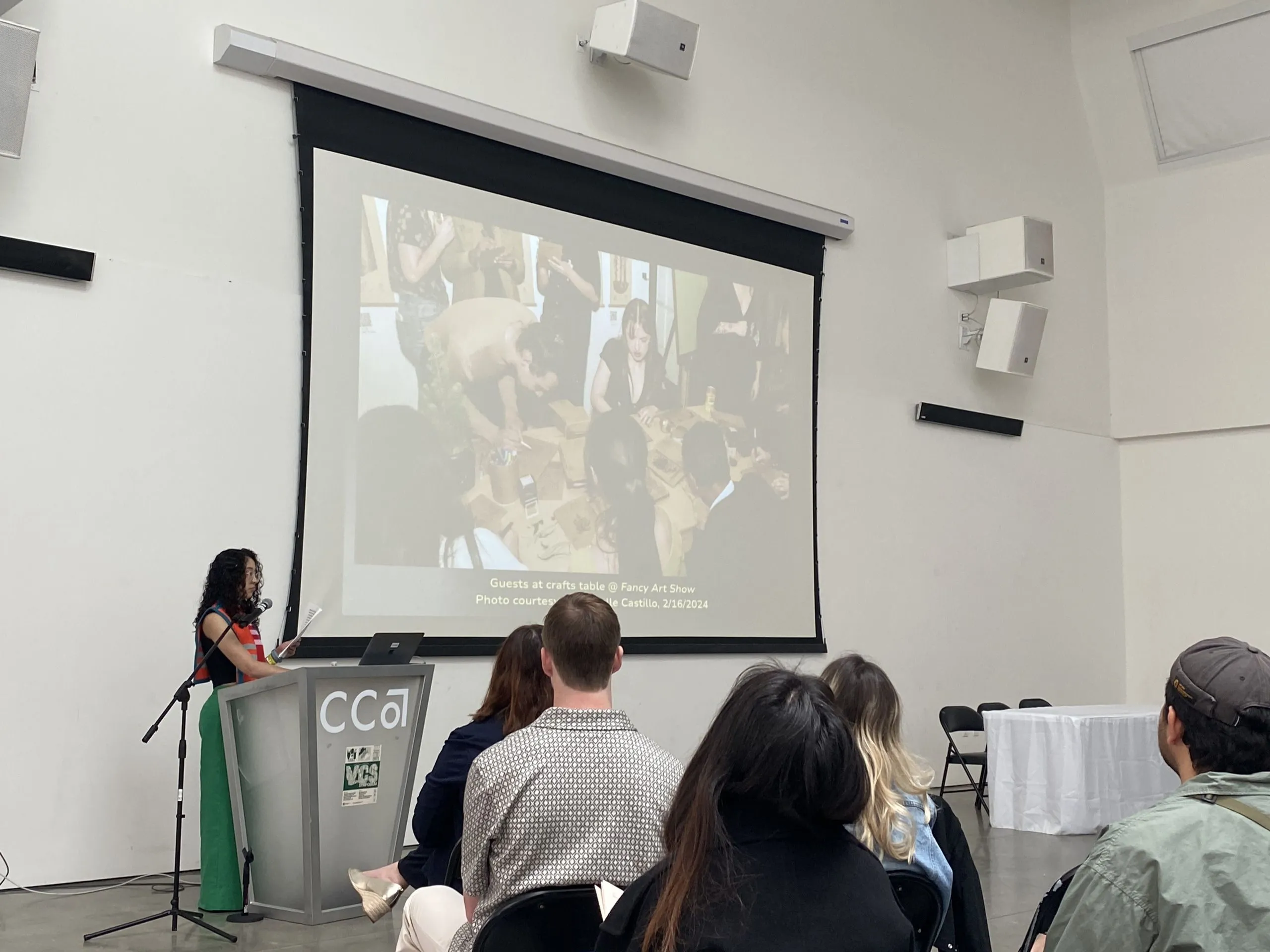 A presenter speaks at a CCA-branded podium during a VCS symposium panel while a projected slide behind her shows attendees gathered at a crafts table.