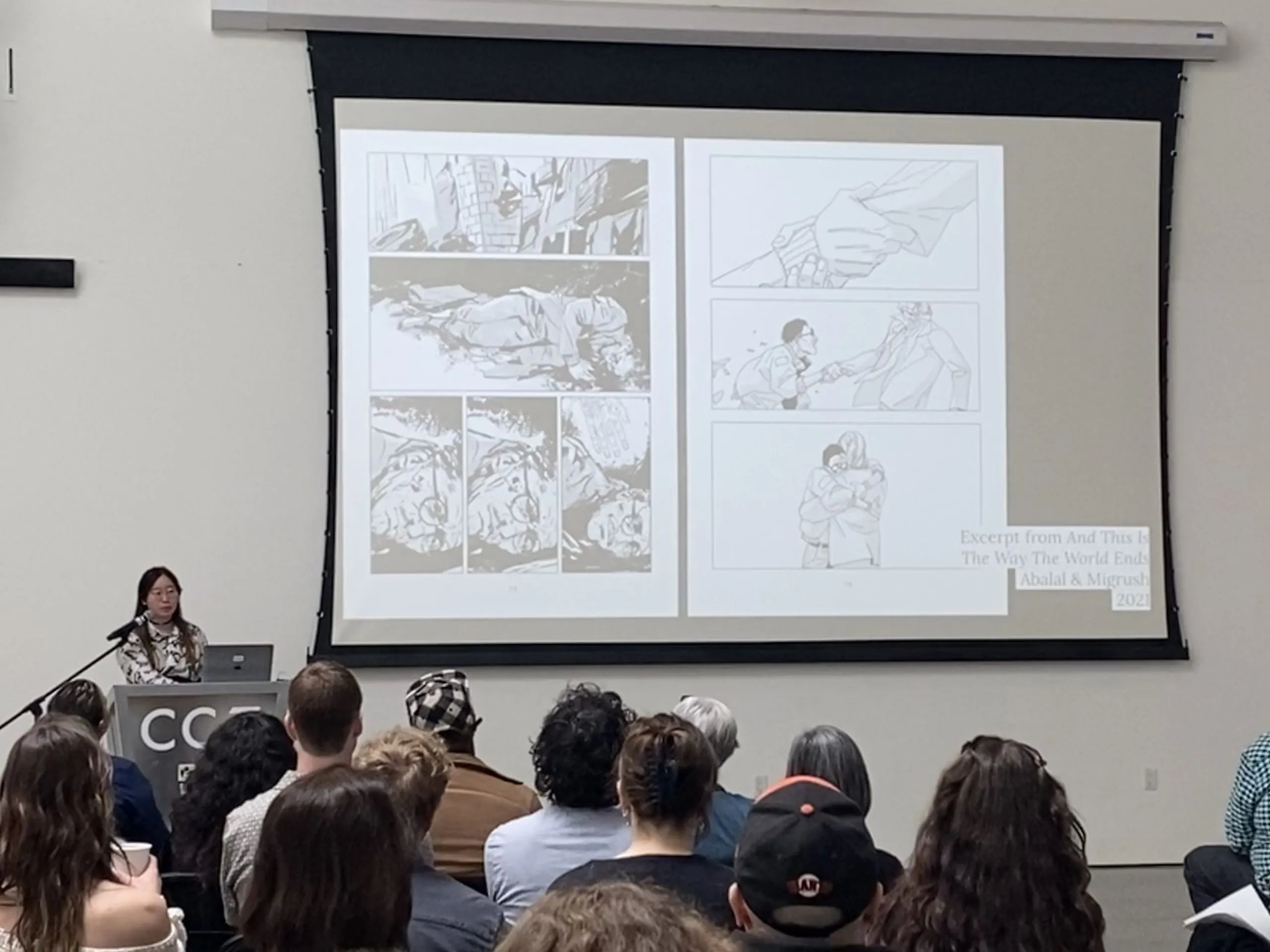A presenter at a podium shows comic book panel artwork on a large projection screen to a seated audience during a VCS symposium talk.