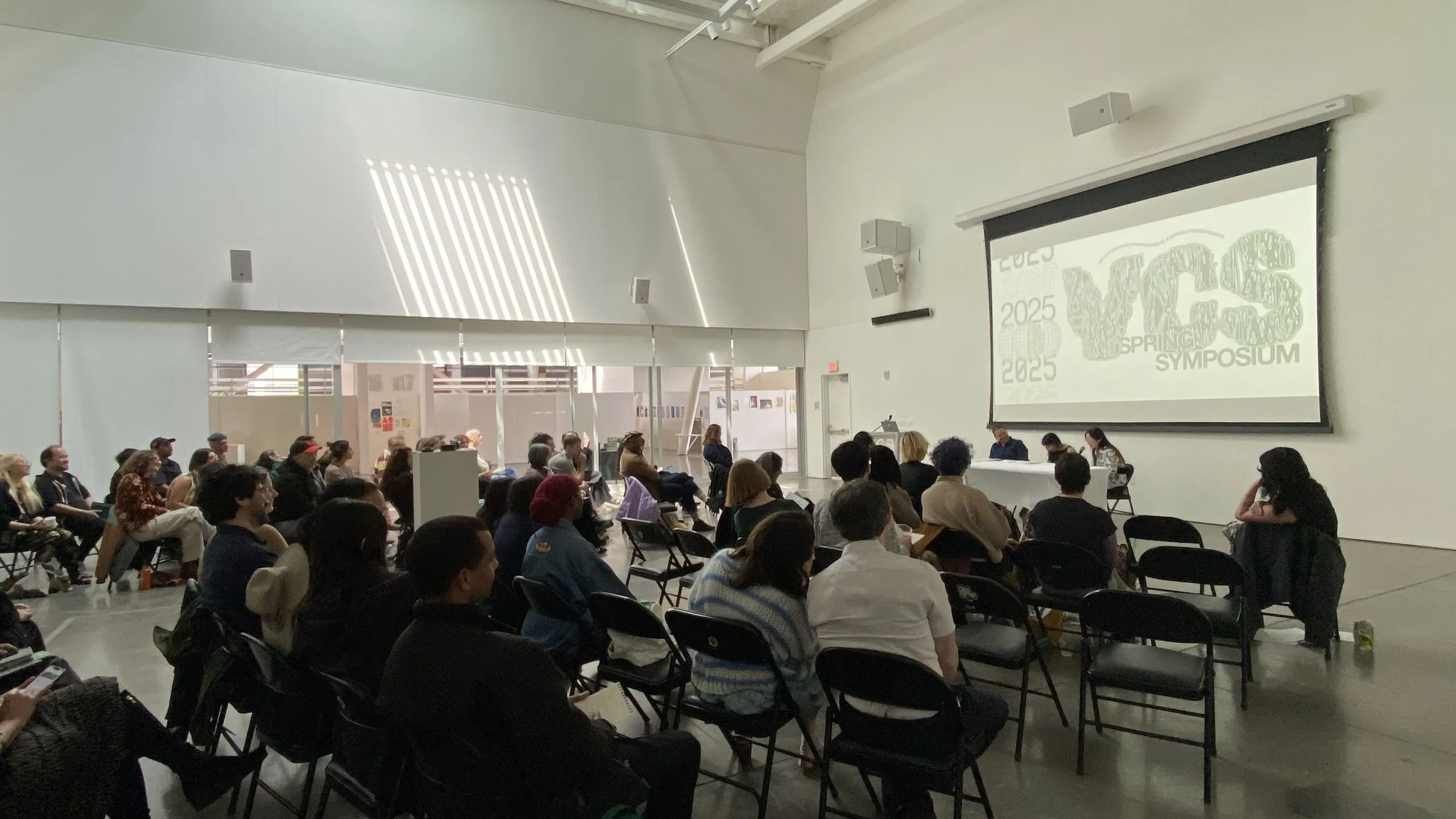 A large audience seated in folding chairs faces a projected VCS Spring Symposium title slide in a bright, white-walled gallery space.