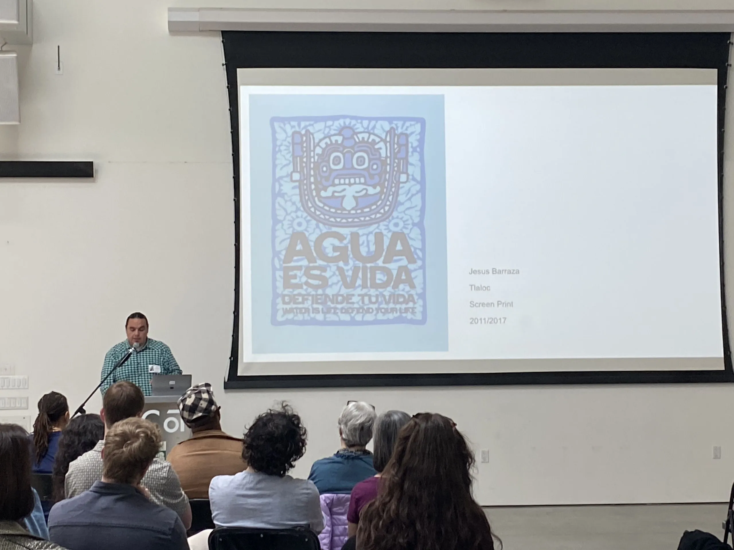 A speaker at a CCA podium presents a blue screen-print reading 'Agua Es Vida' by Jesus Barraza to a seated audience.