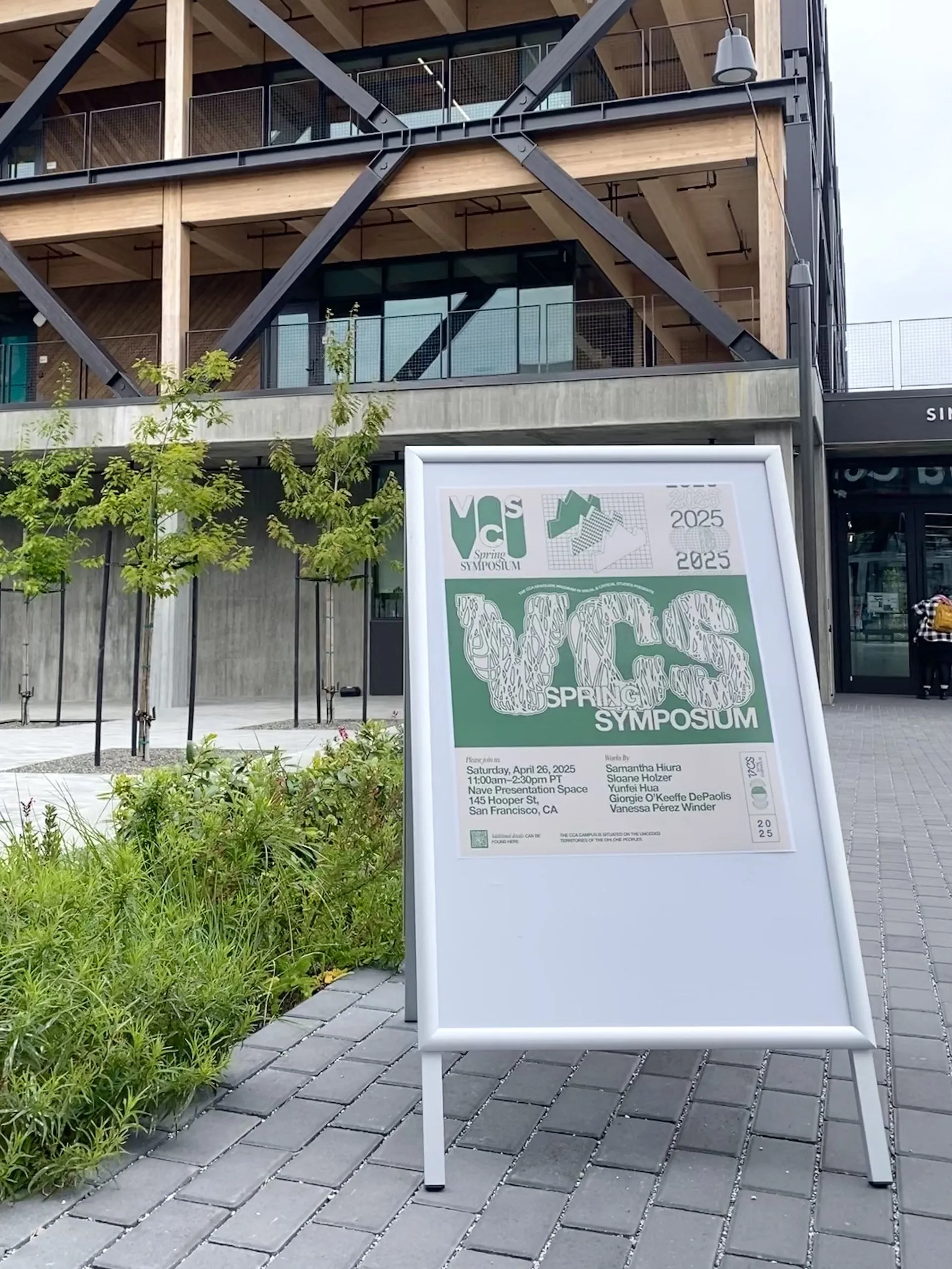 A sandwich board displaying the 2025 VCS Spring Symposium poster stands outside a wood-and-steel CCA campus building.