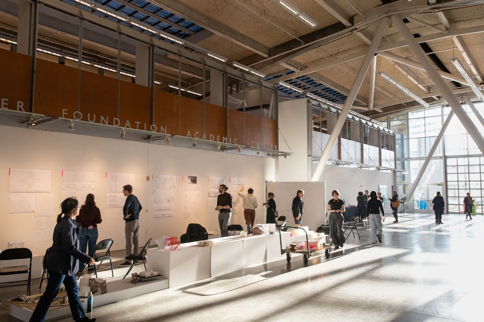Architecture students and reviewers gather in a sunlit studio hall, examining drawings and models pinned along a long white partition wall.