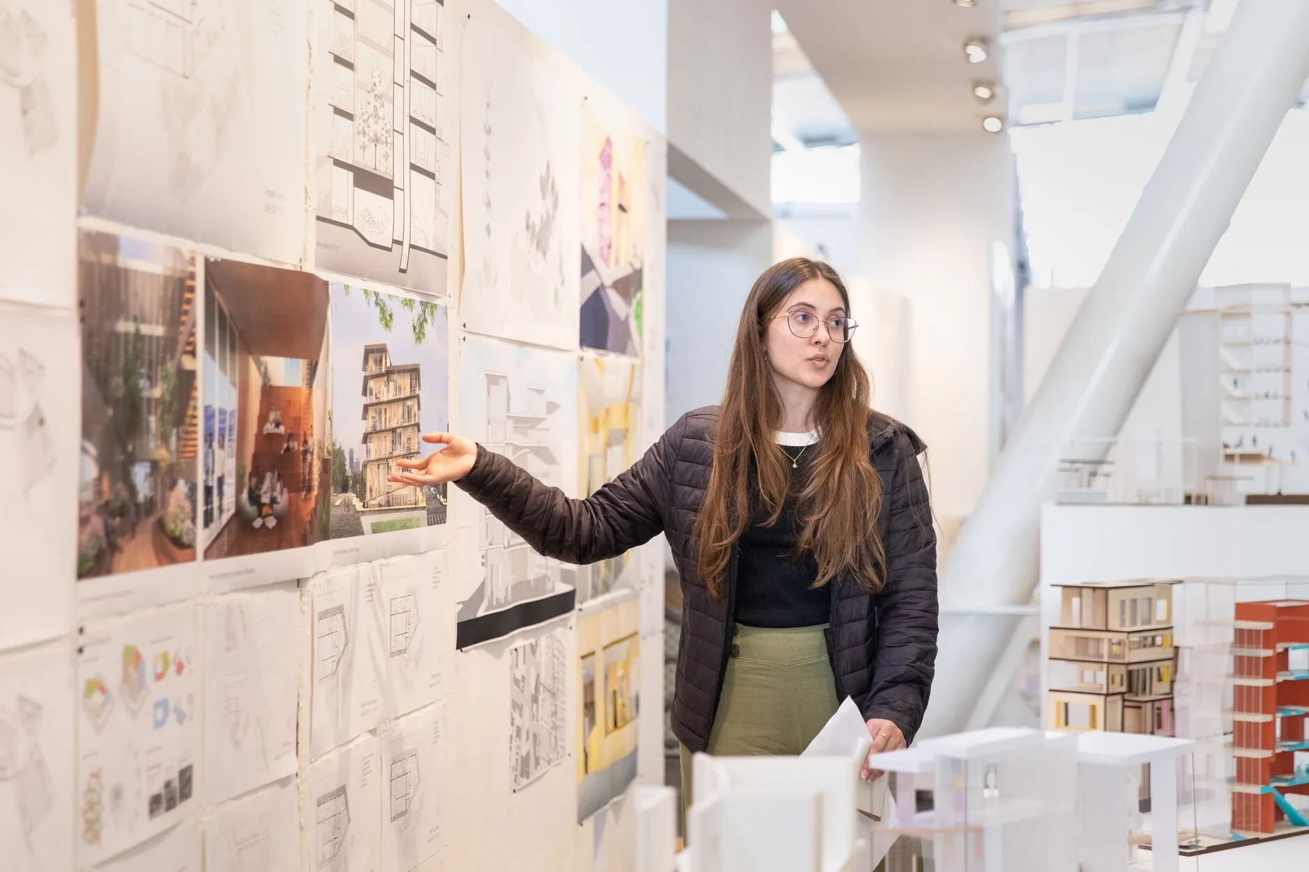 An Architecture student points to pinned renderings and sketches on a critique wall, with white scale models visible on the table beside her.