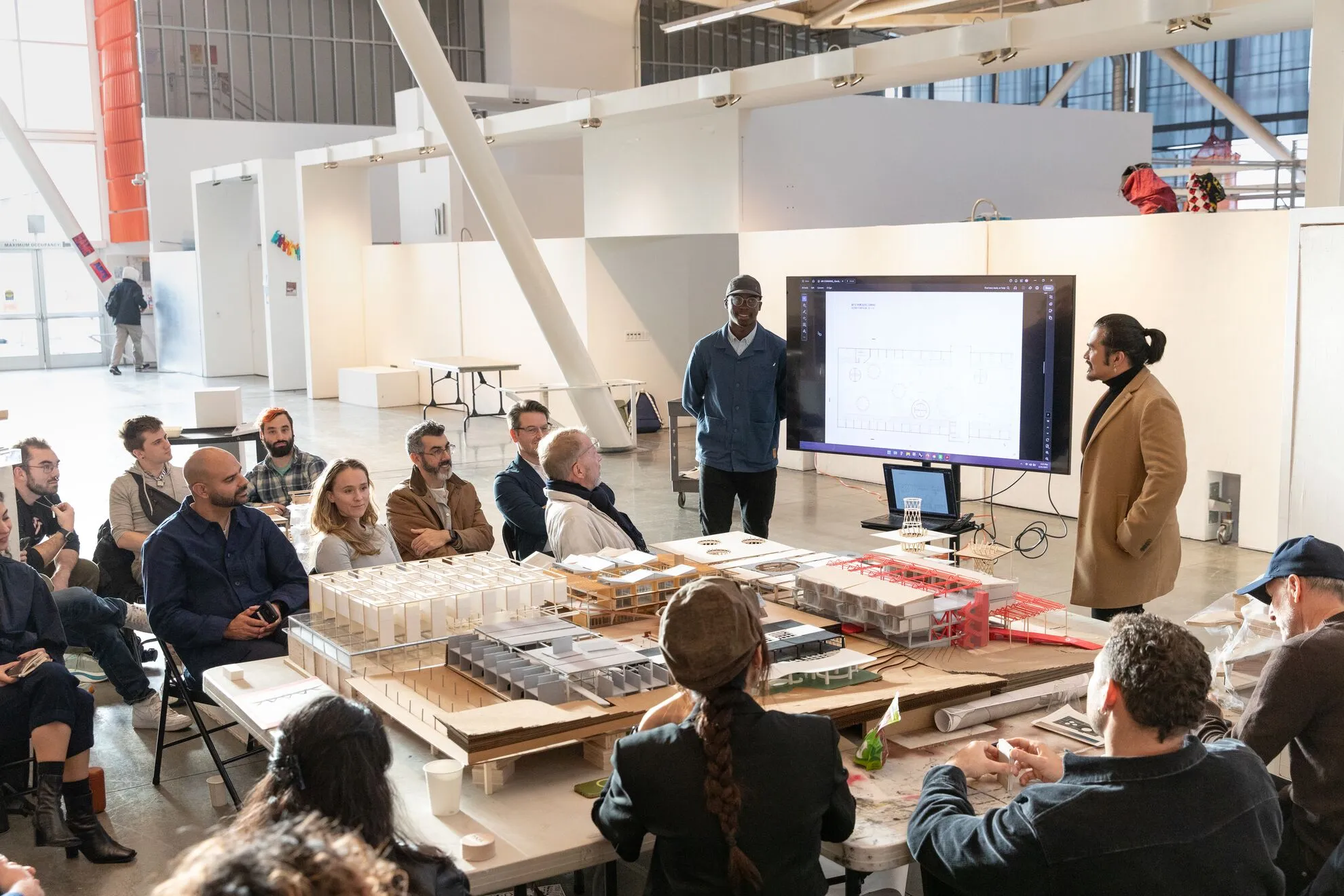 An Architecture student presents to a seated group of critics and peers in a large studio space, with white and red-accented scale models arranged on the table in the foreground.