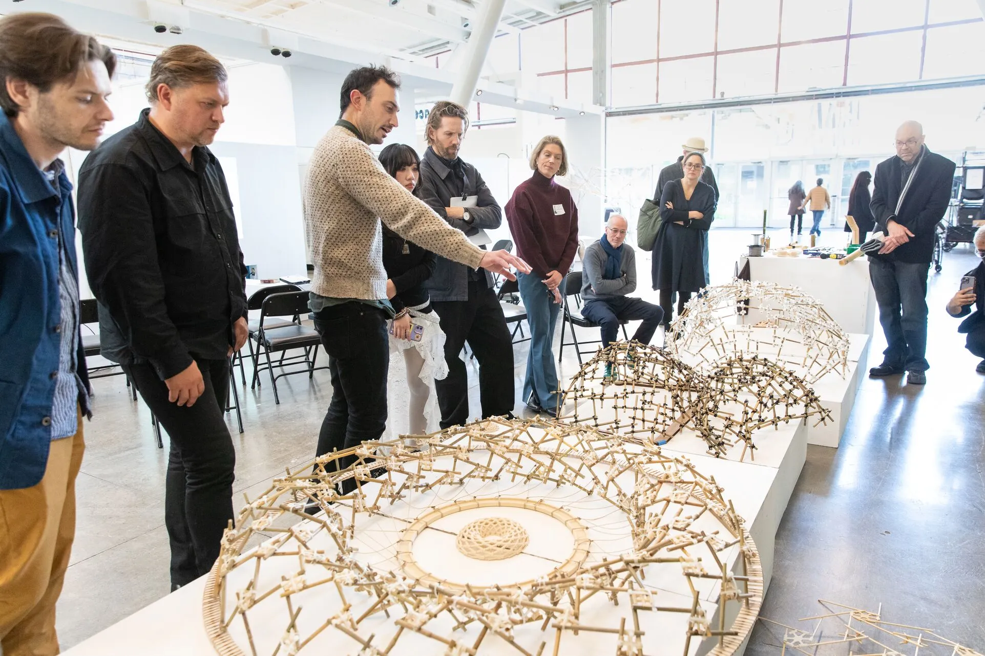 Reviewers and Architecture students lean in to examine a series of large structural models made from thin wooden sticks laid out on a long white table.