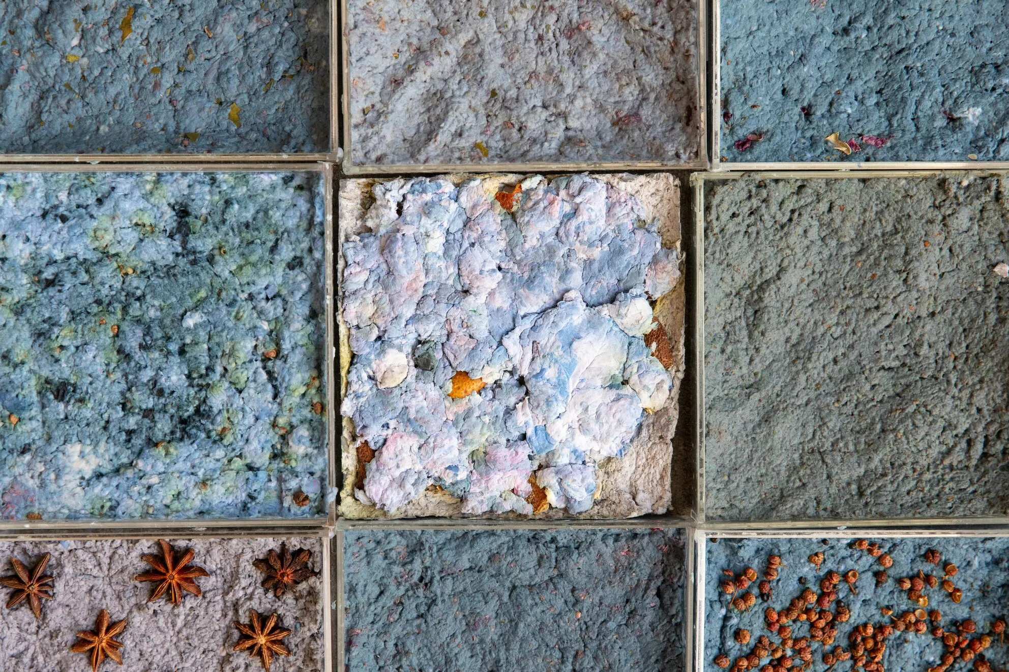 A grid of nine material sample tiles in shades of blue and gray, each embedded with different botanical and granular elements such as star anise and seeds.