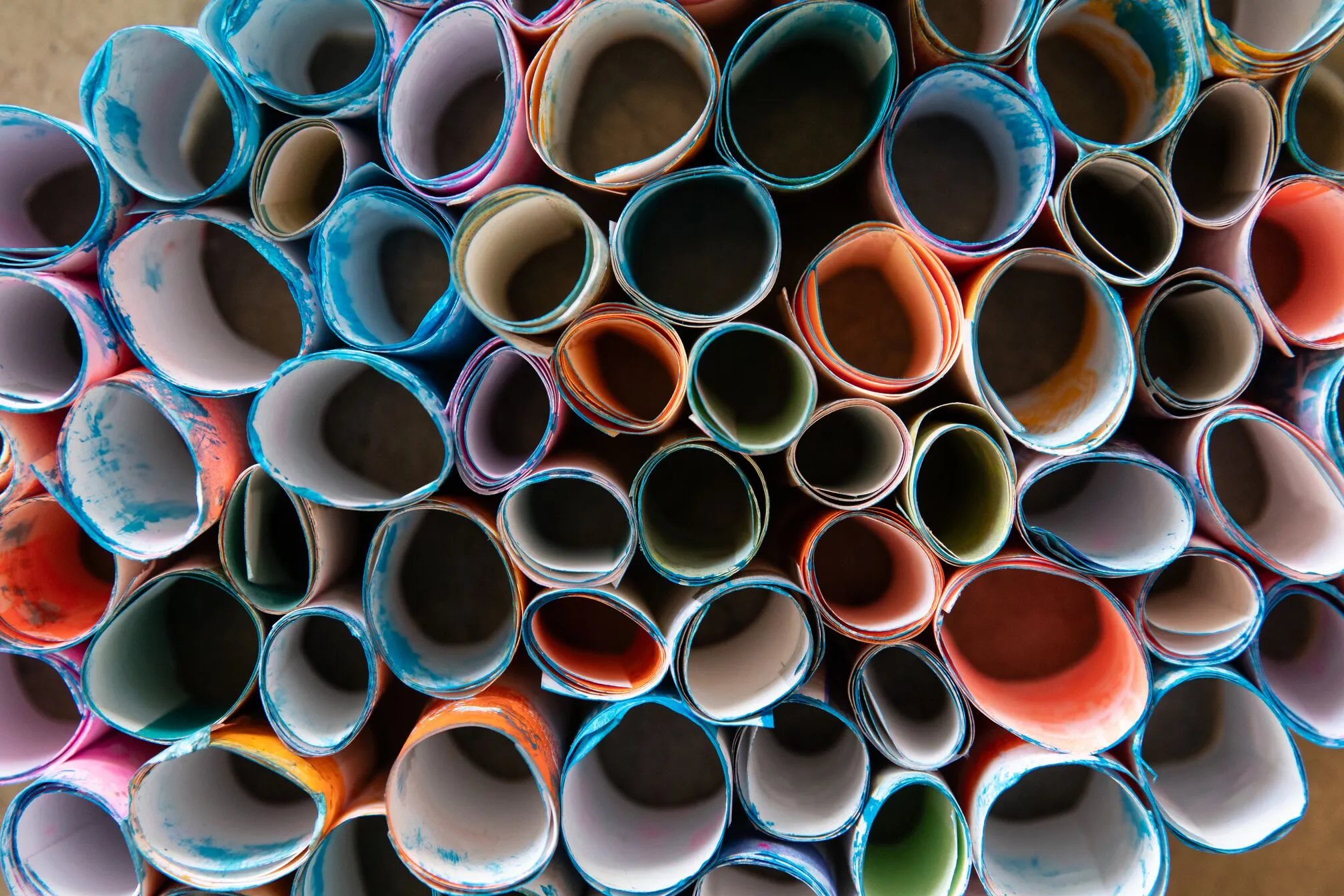 Dozens of tightly rolled paper sheets in mixed colors are stacked end-on, revealing circular cross-sections of blue, red, orange, and green painted edges.