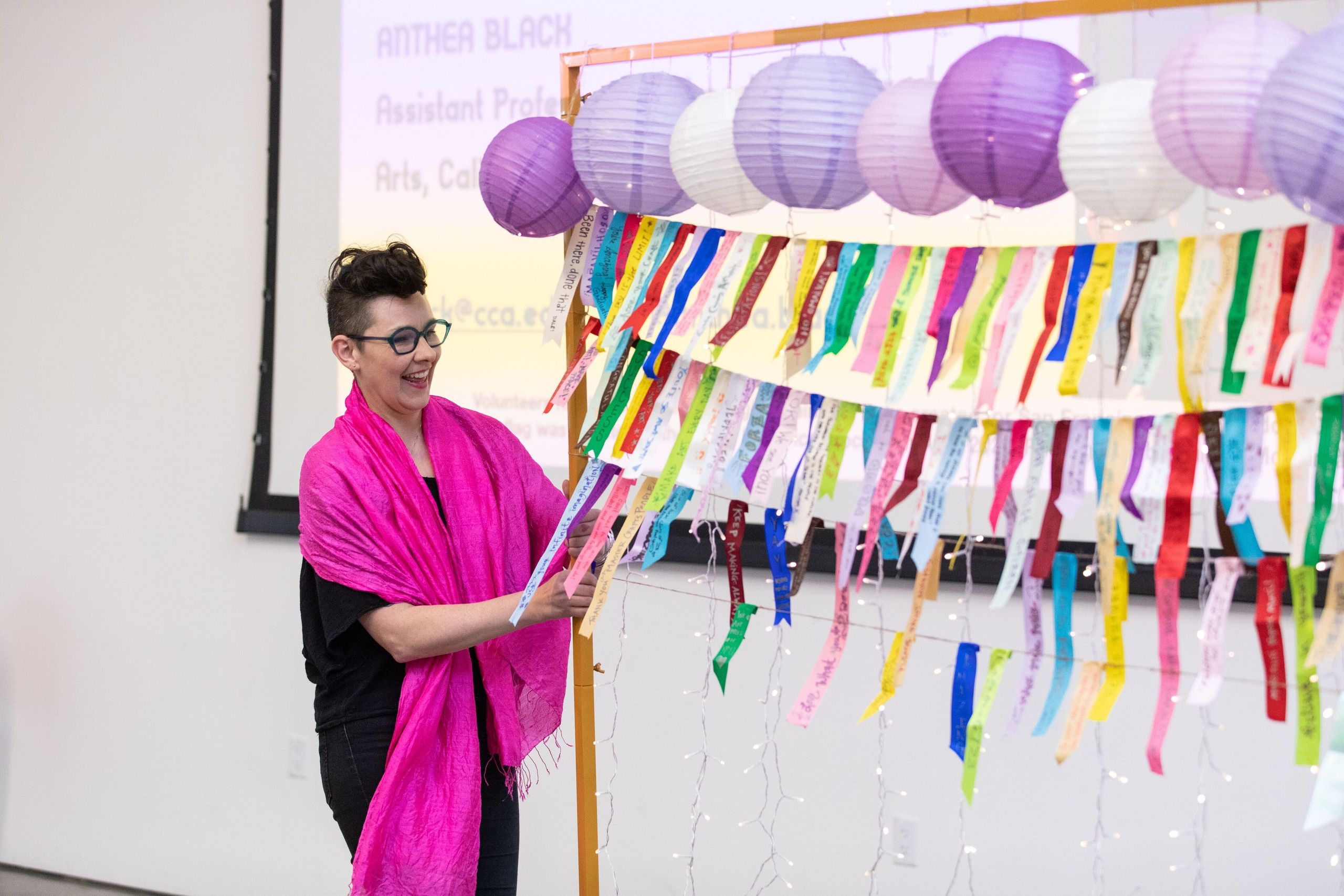 A graduate in a vibrant pink shawl stands next to colorful paper lanterns and rainbow streamers, smiling during a commencement celebration.