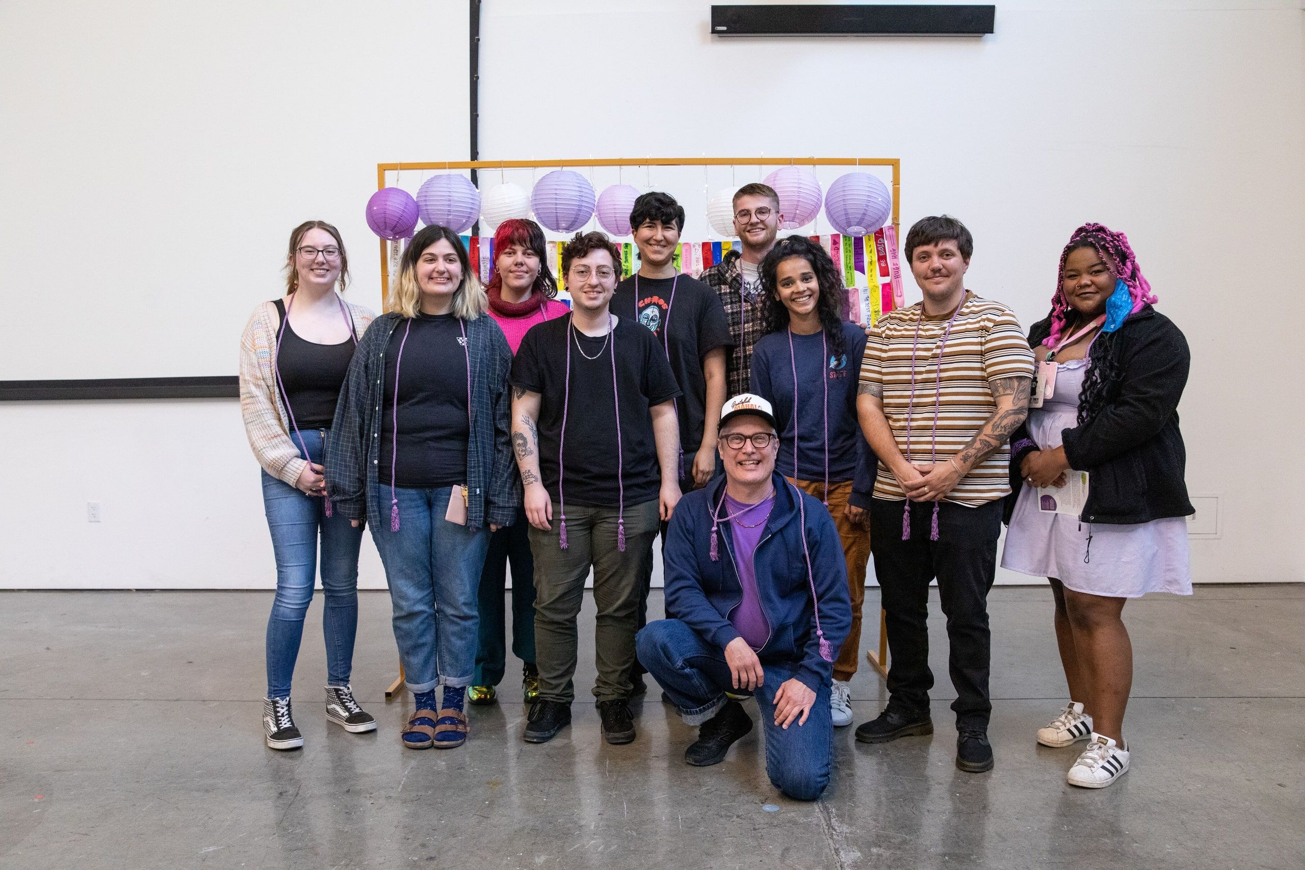 A group of ten students dressed in casual clothing pose together in front of decorative purple lanterns and colorful ribbons at an indoor celebration.