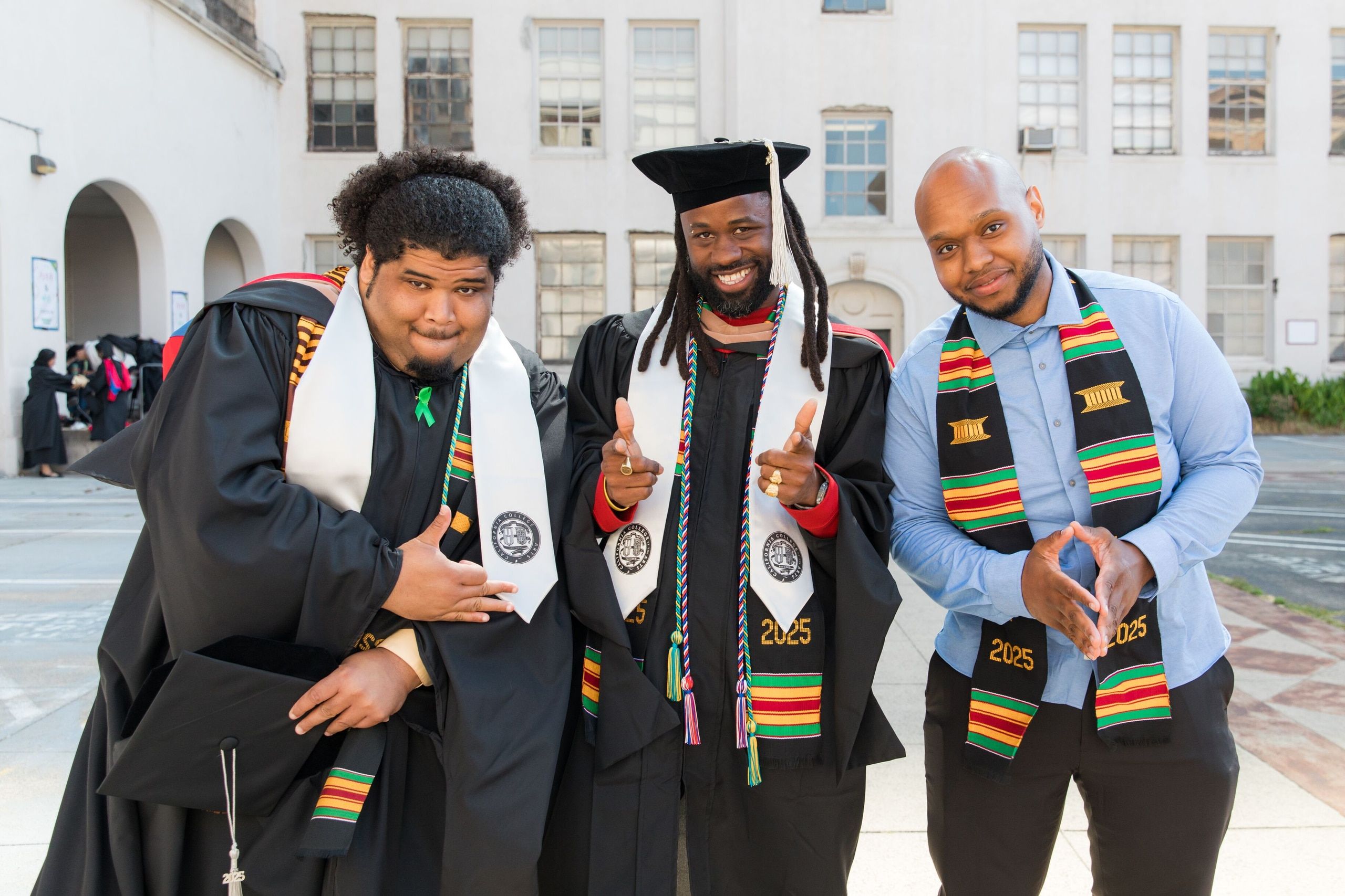 Three male graduates wearing black robes and colorful cultural sashes stand together outdoors on a brick plaza, smiling at the camera.