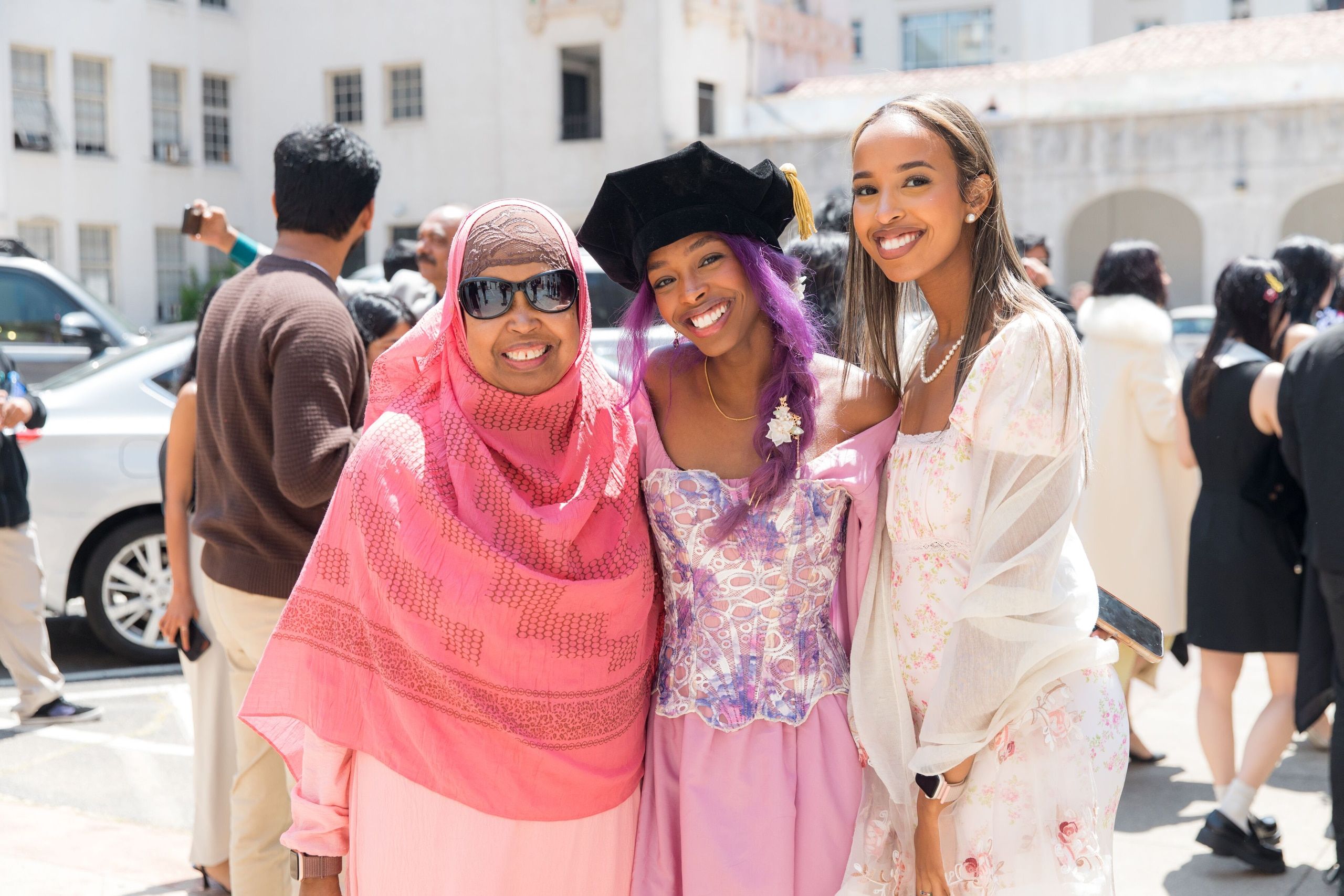 Four graduates dressed in vibrant pink and purple outfits pose together in a parking lot during a bright, sunny commencement day.