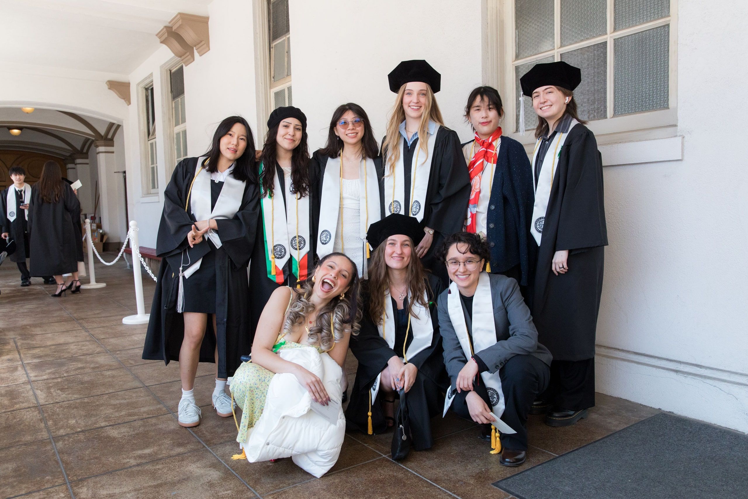 Nine graduates in black academic robes and regalia pose together indoors, with two seated in front and seven standing behind them.