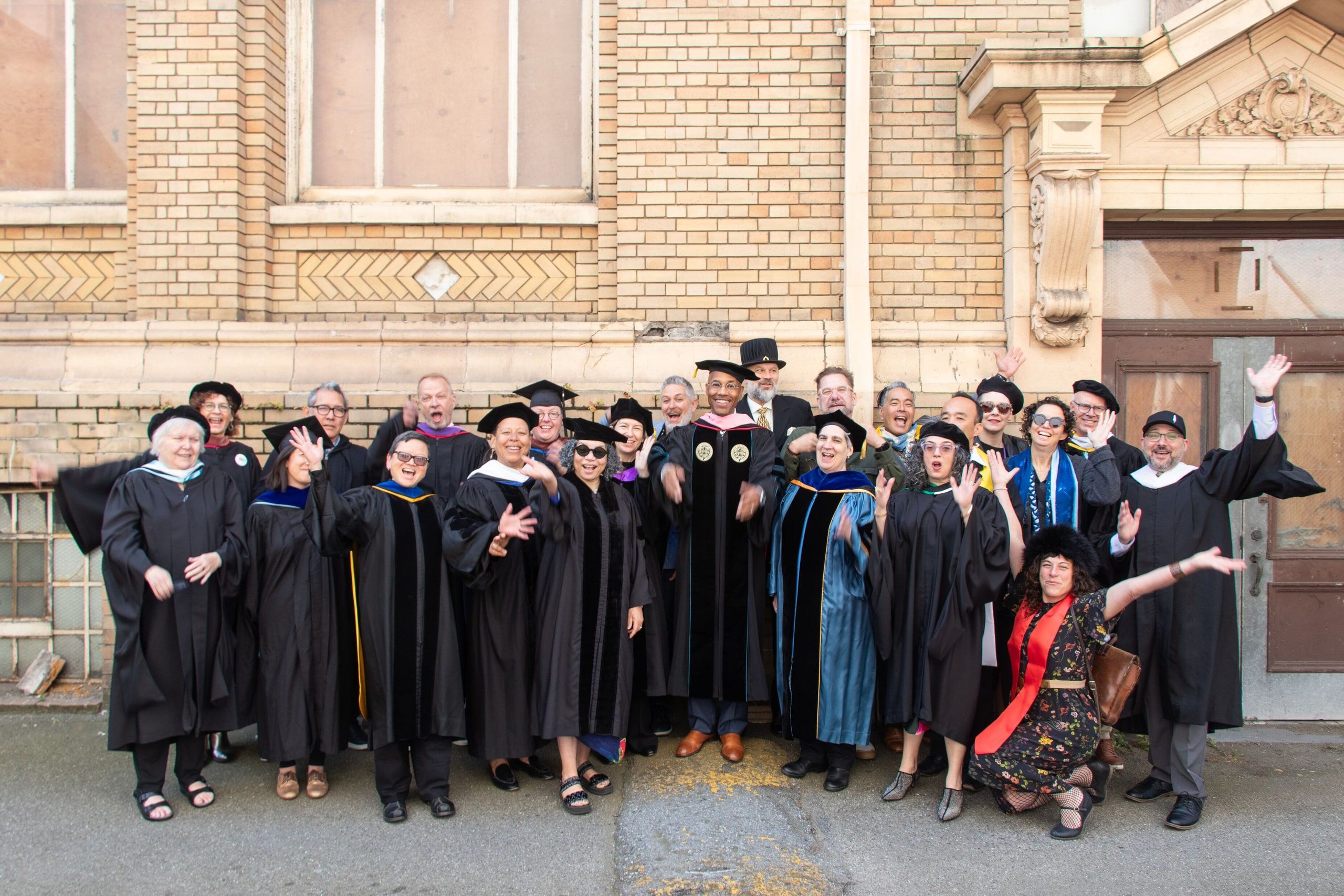 A group of graduates in black robes and mortarboards stand together in front of a brick building, celebrating with raised arms and smiling at the camera.