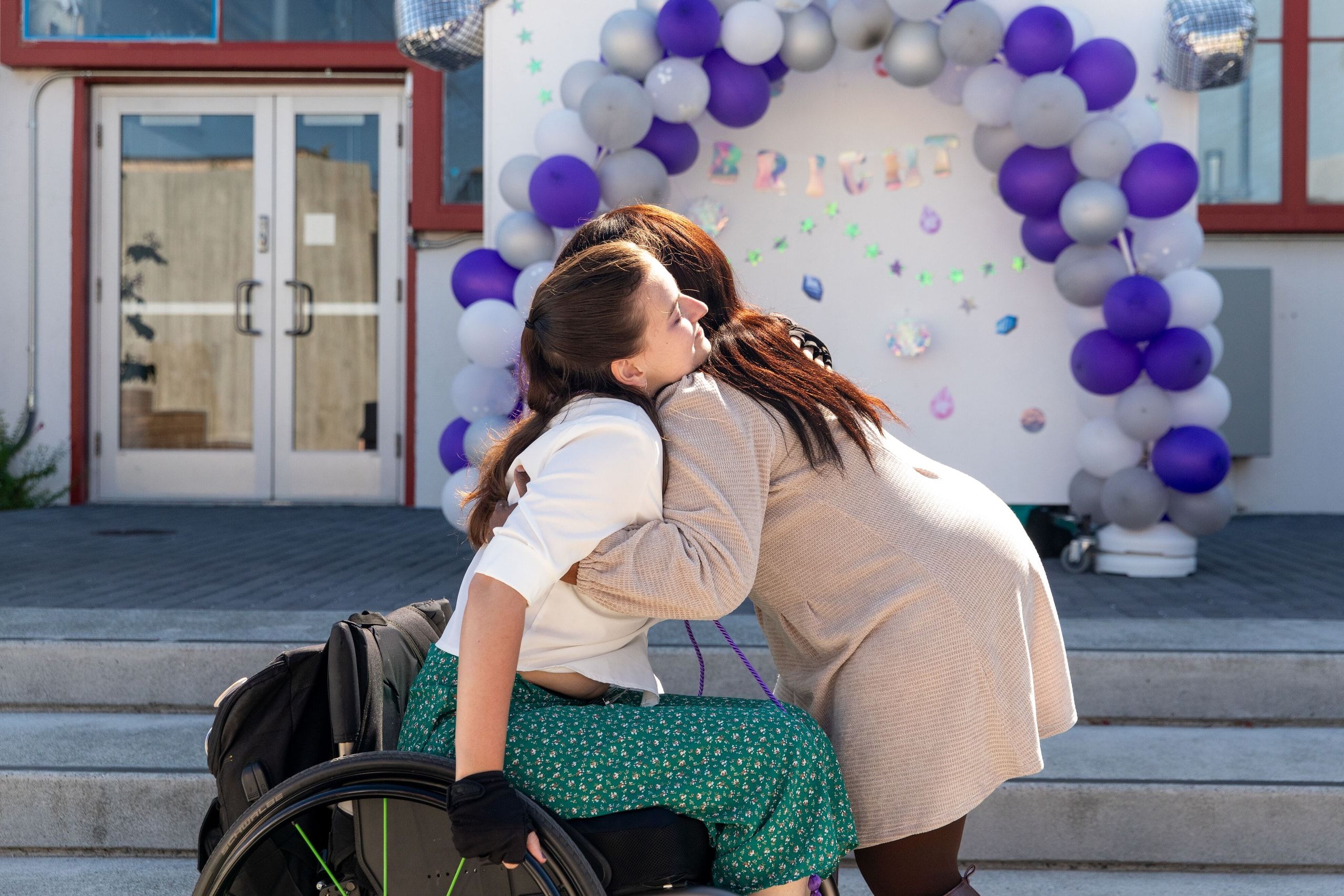 Two graduates embrace in front of a celebration backdrop decorated with purple and white balloons, with a paved courtyard and building visible behind them.