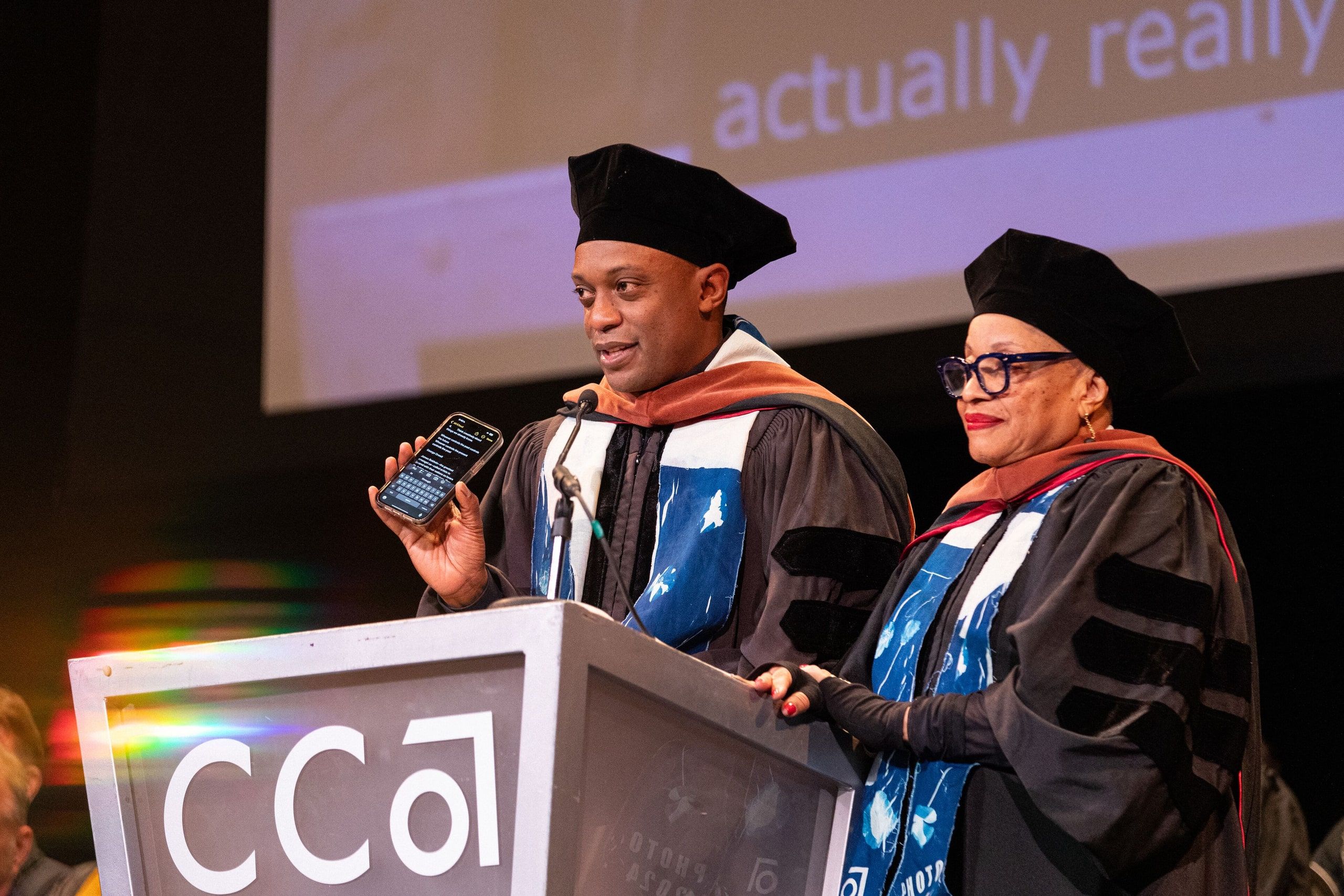 Two speakers in academic regalia stand at a CCAlum podium during a commencement ceremony, with one holding a microphone as they address the audience.