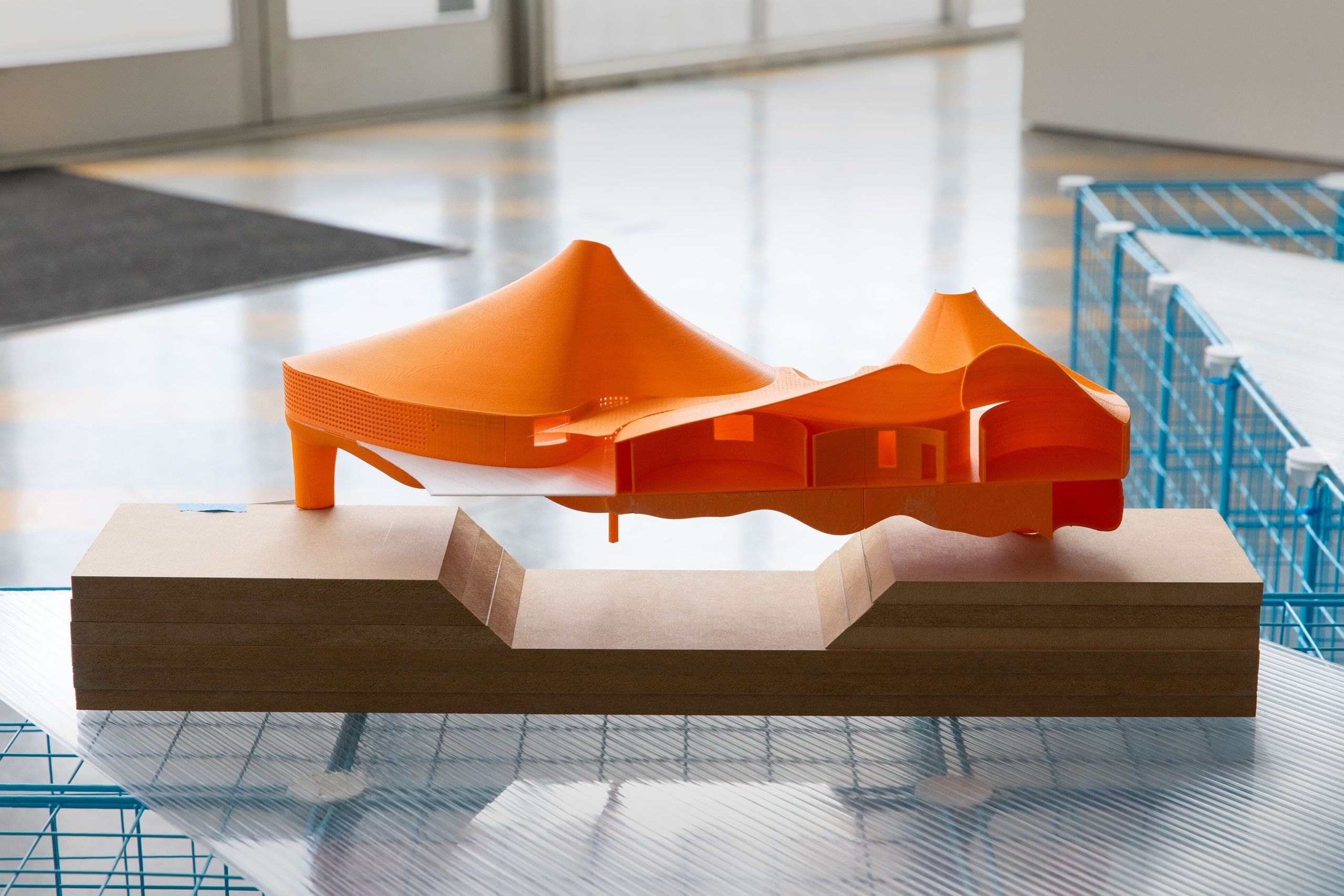 A bright orange sculptural model with curved and geometric forms sits on a light wood base in a gallery space with polished floors.