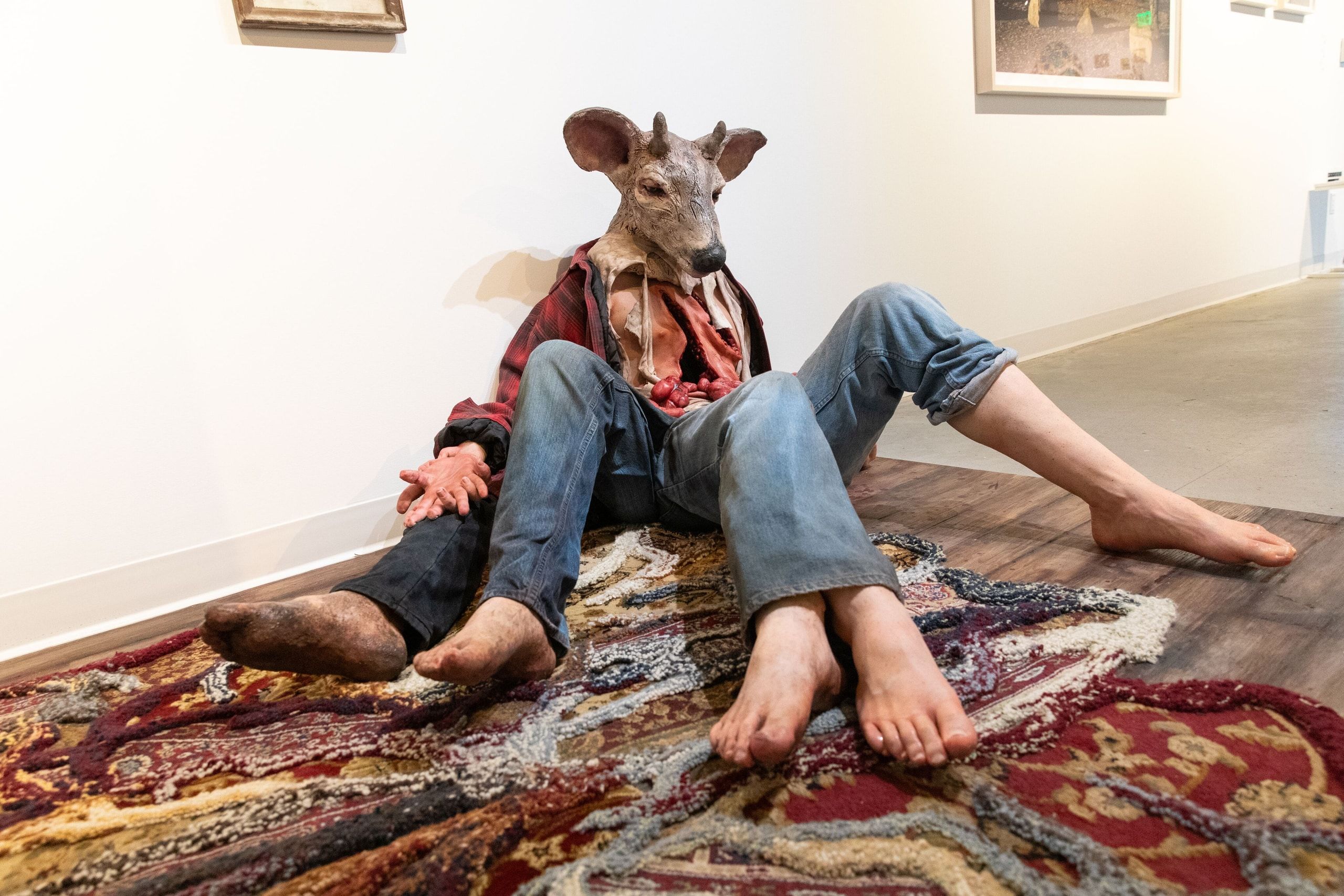 An art installation featuring a figure wearing a deer head mask seated on a patterned textile floor, creating a surreal and contemplative composition.