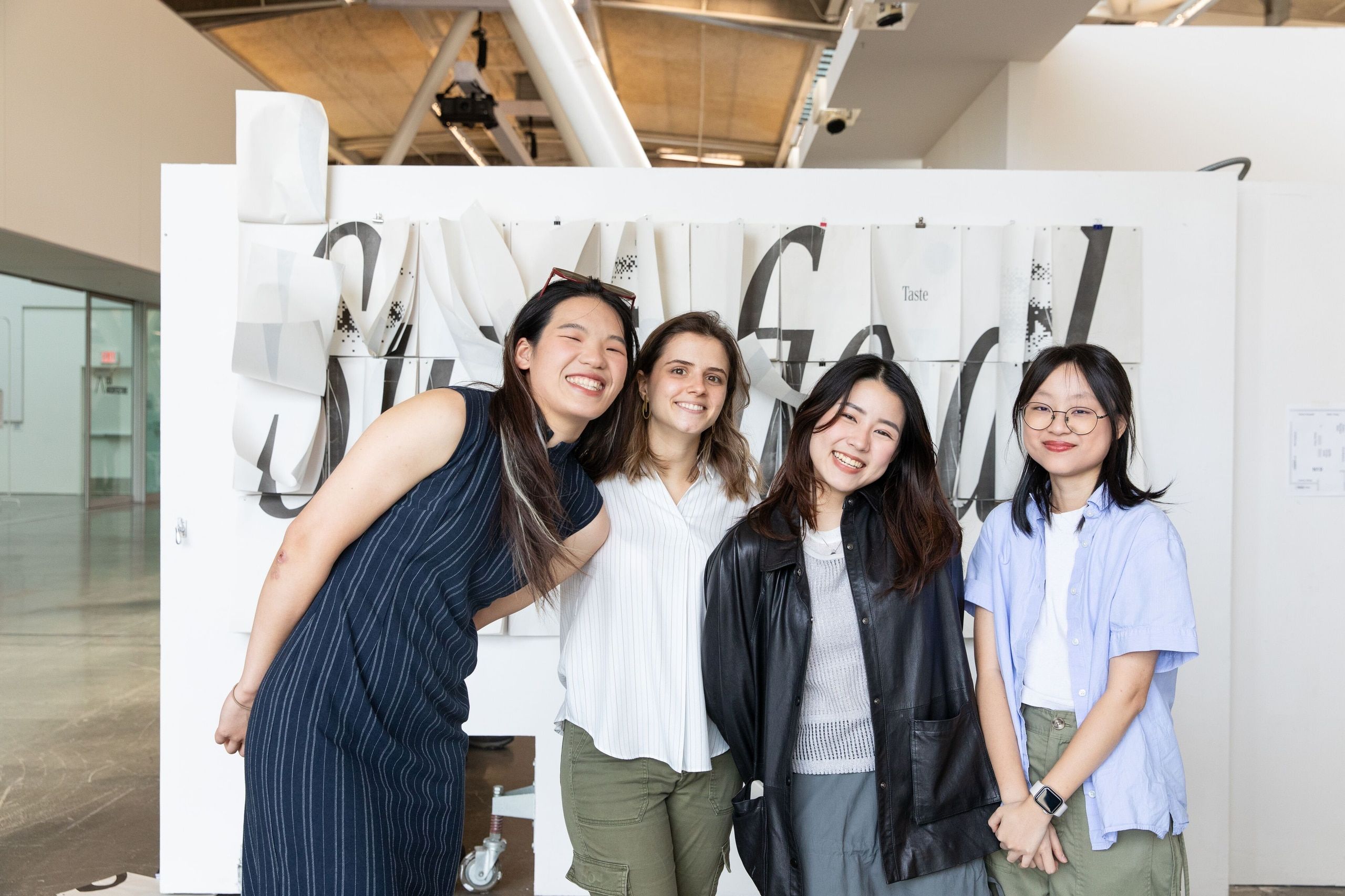 Four student artists pose together in front of their large-scale white exhibition display in a gallery space.