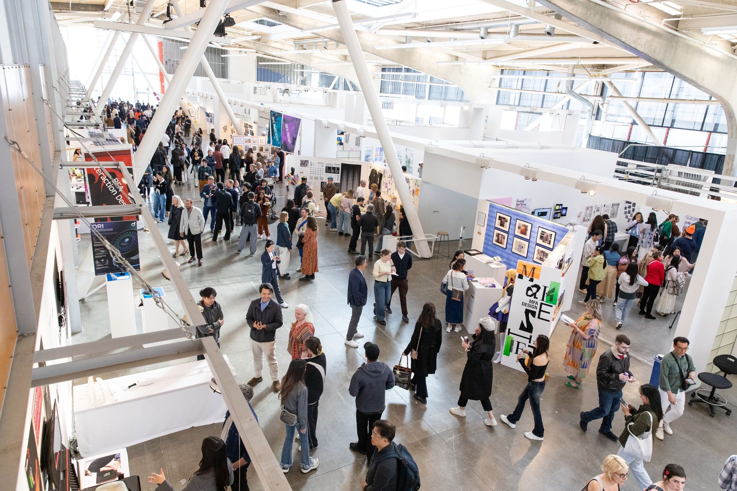 Overhead view of a bustling gallery space with multiple levels showing numerous visitors viewing diverse student artwork installations and displays.