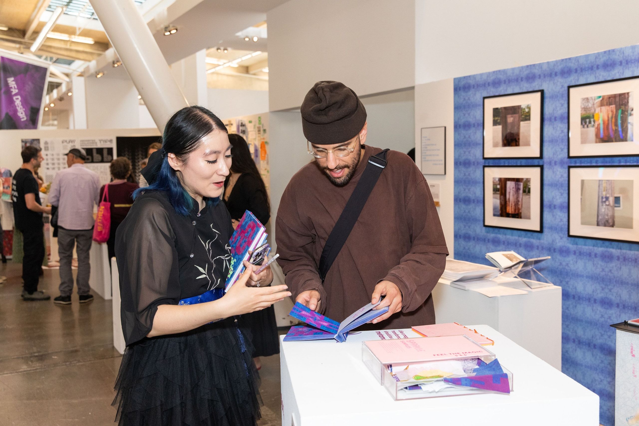 Two visitors examine colorful printed materials and publications at a gallery display table with framed photographs visible on a blue wall panel behind them.