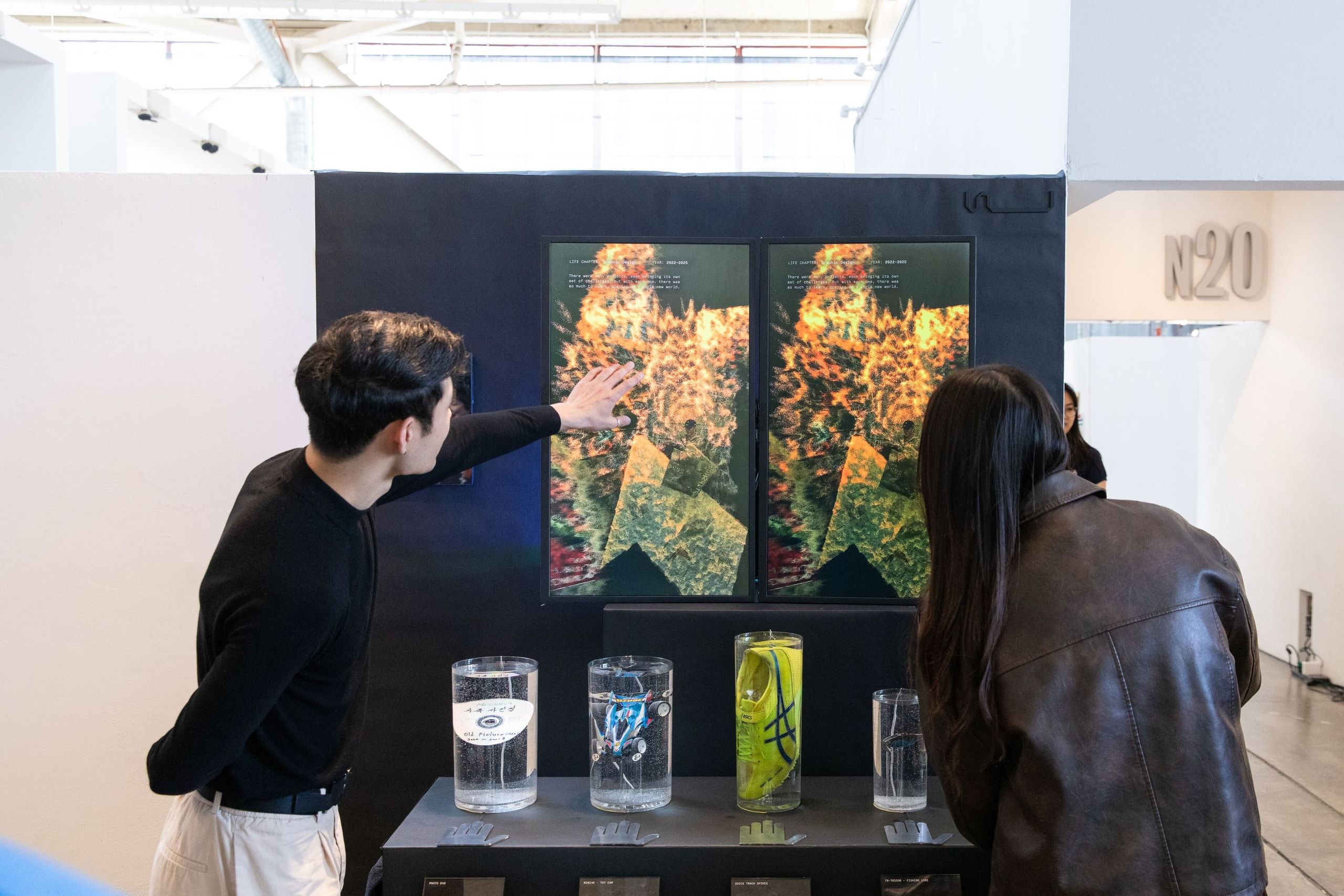 Two visitors view a digital display showing vibrant floral imagery mounted on a dark wall, with water glasses placed on a shelf below.