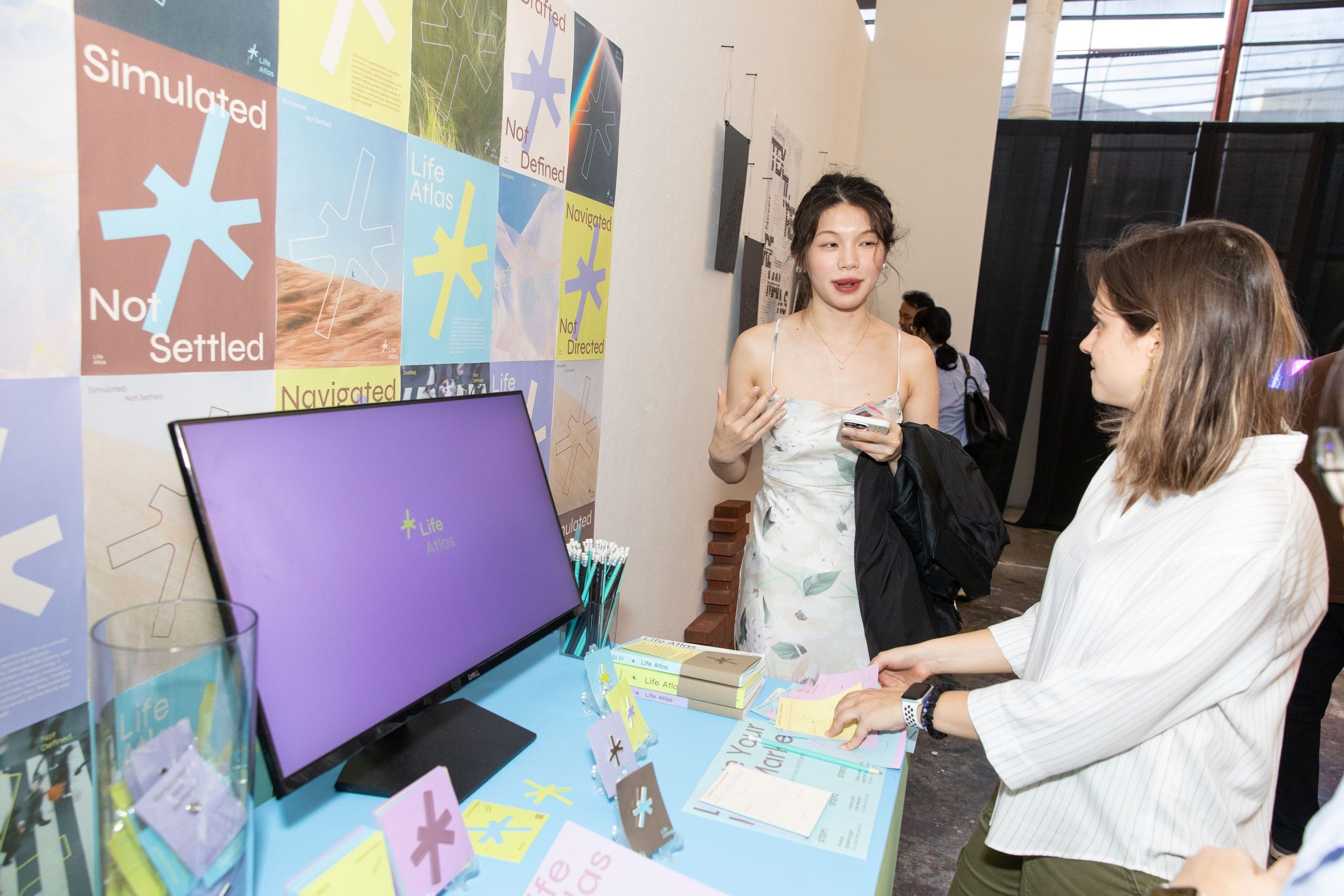 A student artist presents her colorful digital project at a gallery booth, with vibrant yellow, blue, and coral colored posters and signage visible in the background.