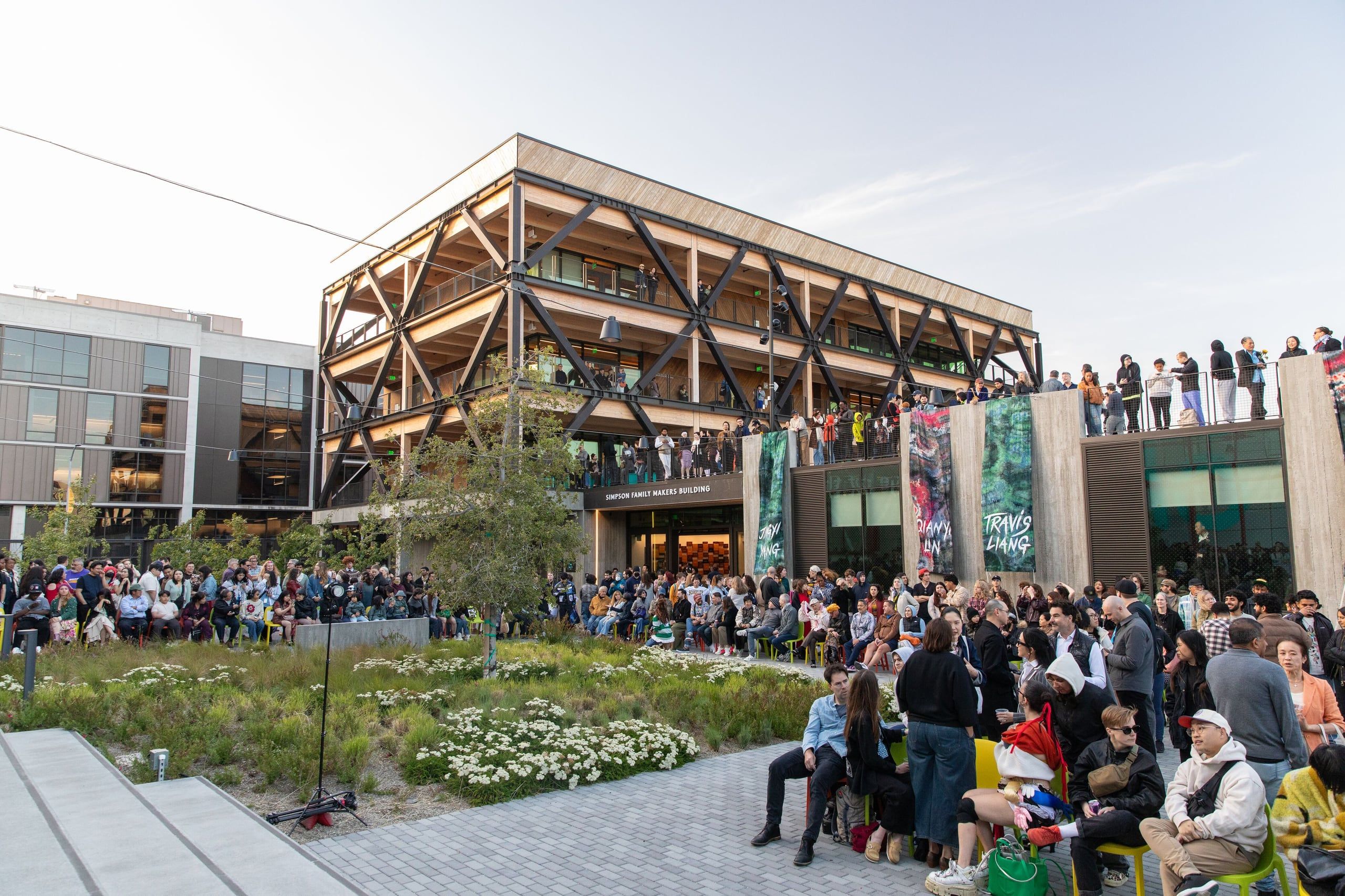 A large crowd of visitors gathers outside a contemporary building with distinctive wooden architectural framework and white flowers in the landscaping.