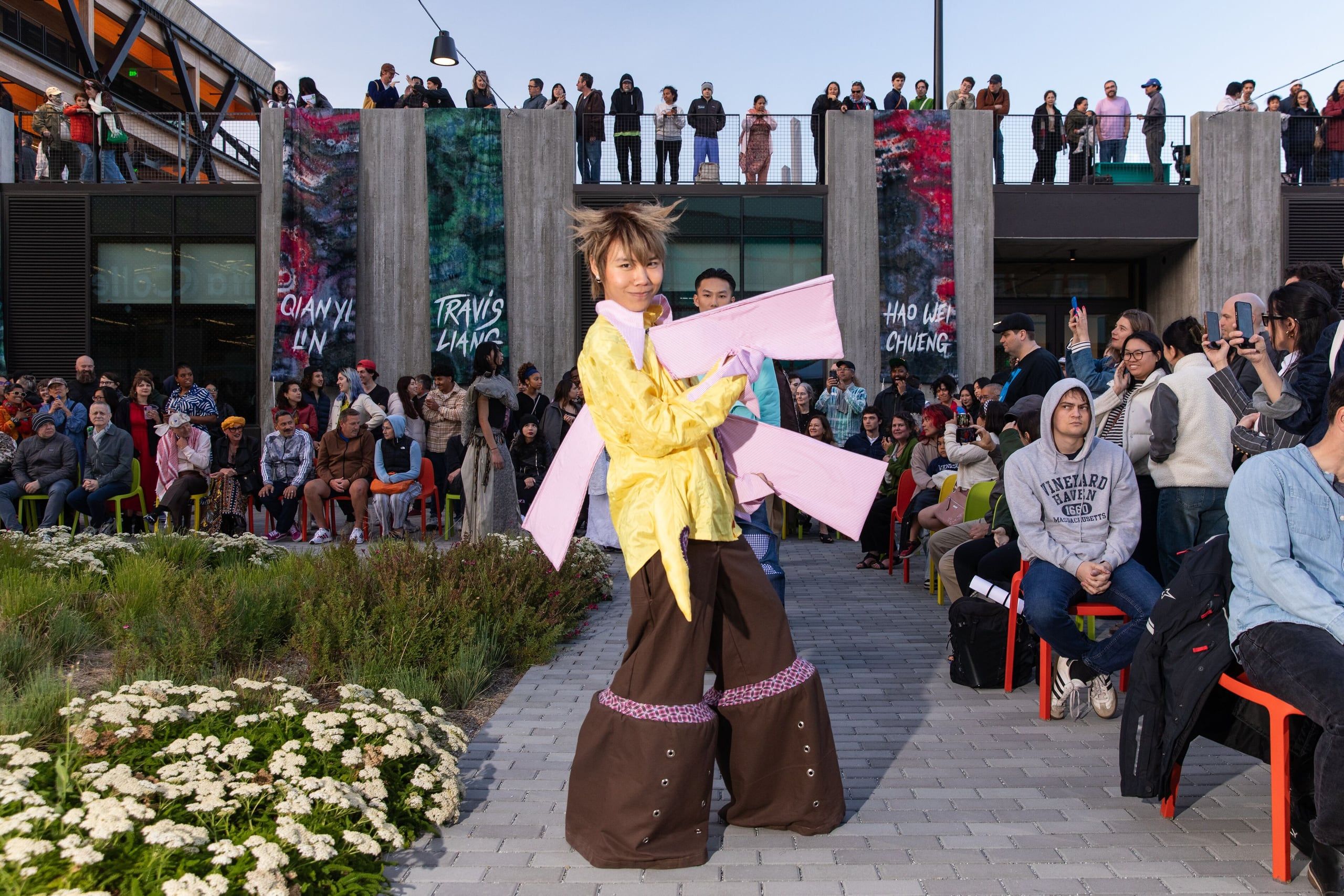 A performer wearing a bright yellow jacket and pink wings stands on a paved outdoor plaza surrounded by spectators and tall wooden pillars with colorful artwork.