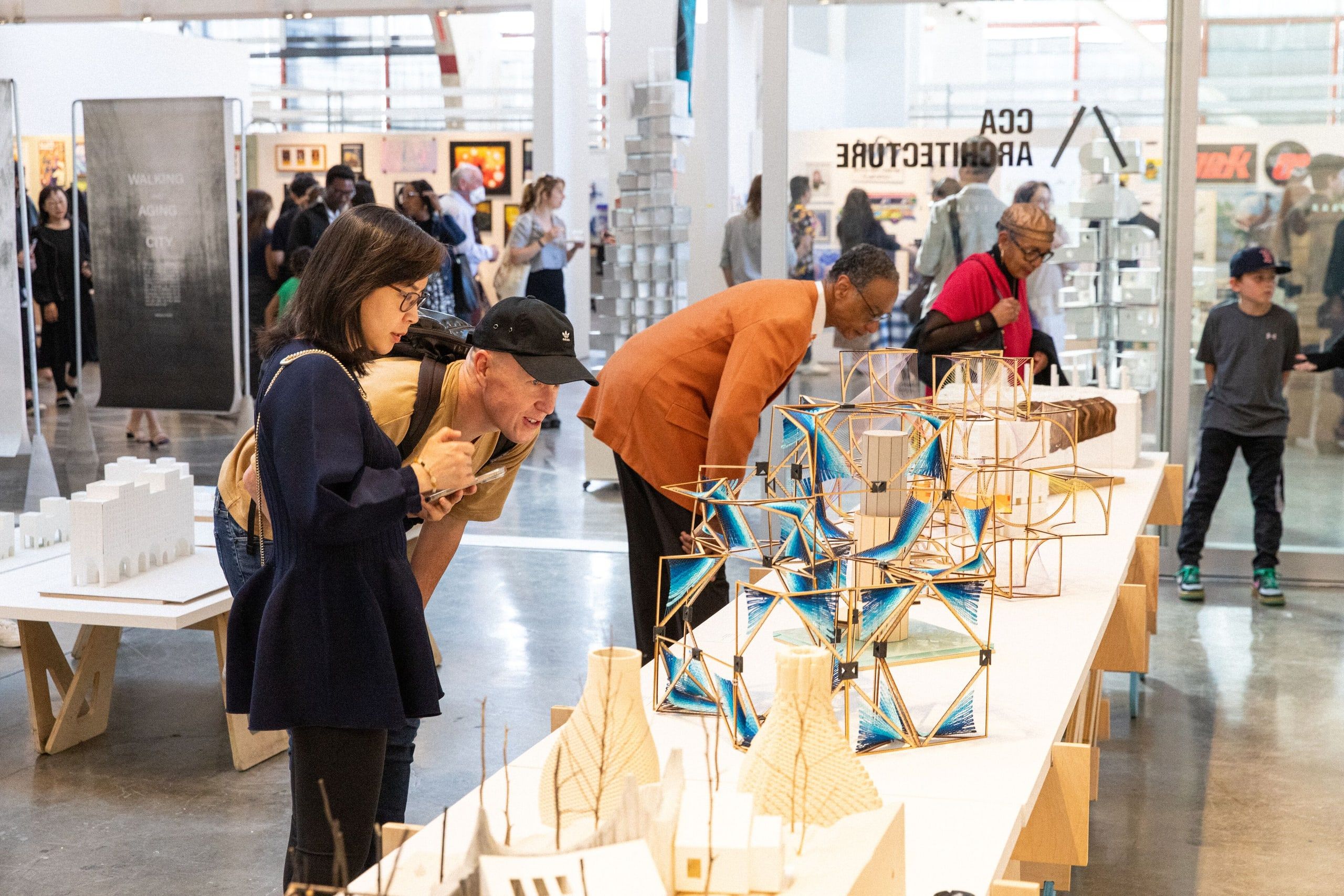 Visitors examine a gallery space featuring wooden geometric sculptures and architectural models displayed on white pedestals.