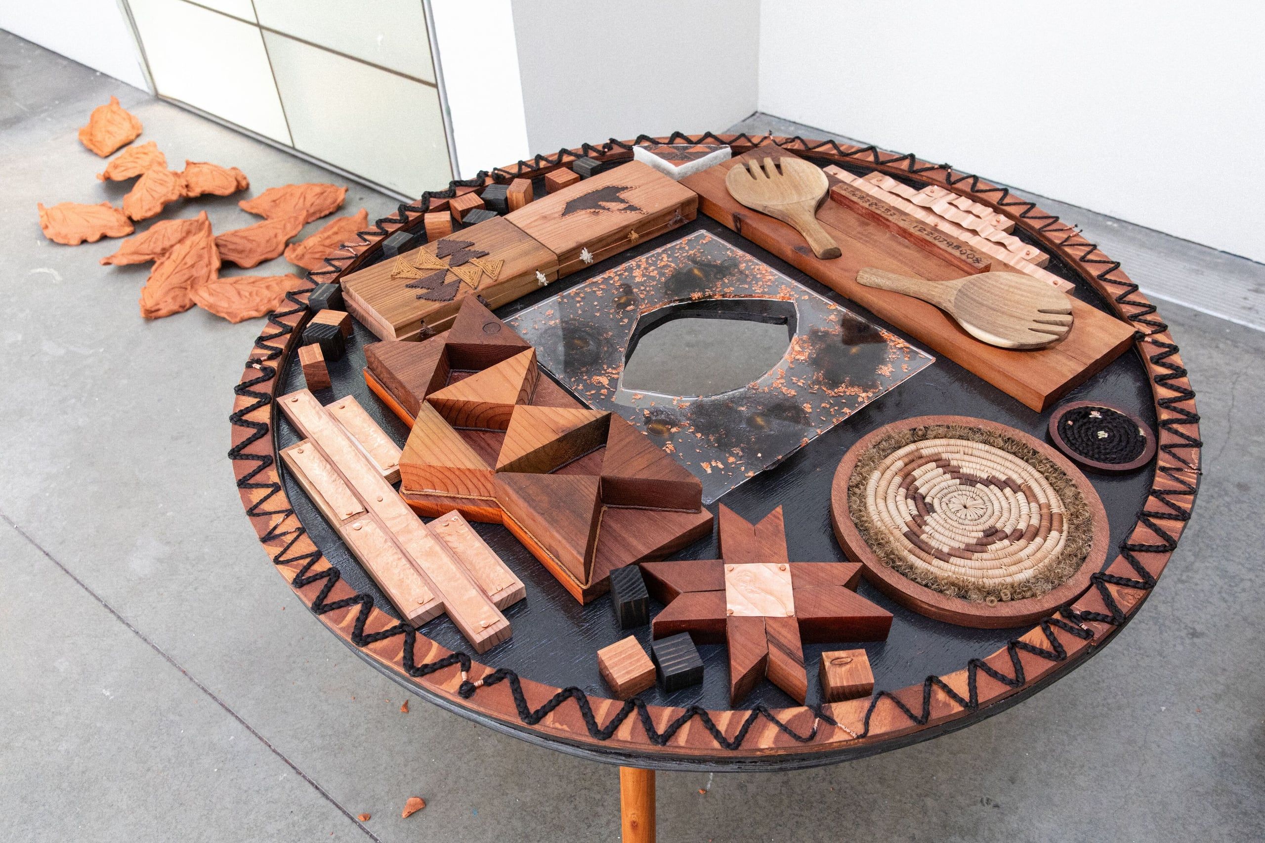 A large circular wooden sculptural installation features geometric patterns, carved elements, and various hand-crafted wooden pieces arranged in a mandala-like composition.