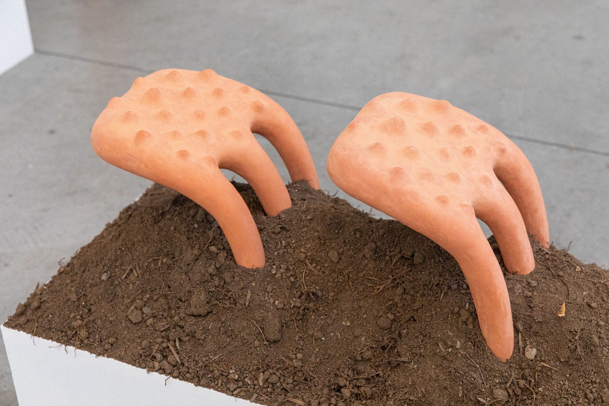 Two terracotta sculptural hands with textured surfaces emerge from a mound of dark soil on a gallery floor.