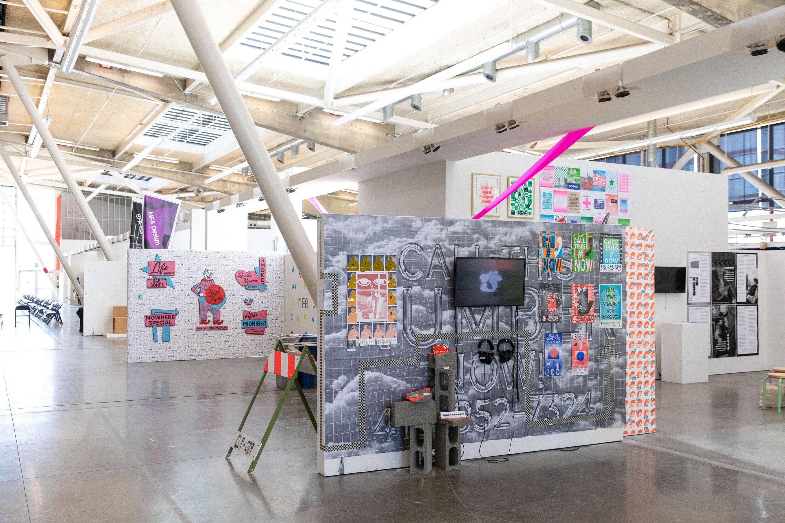 A bright gallery space with industrial ceilings displays multiple large-scale installations including colorful panels and a dark sculptural structure with mixed media elements.