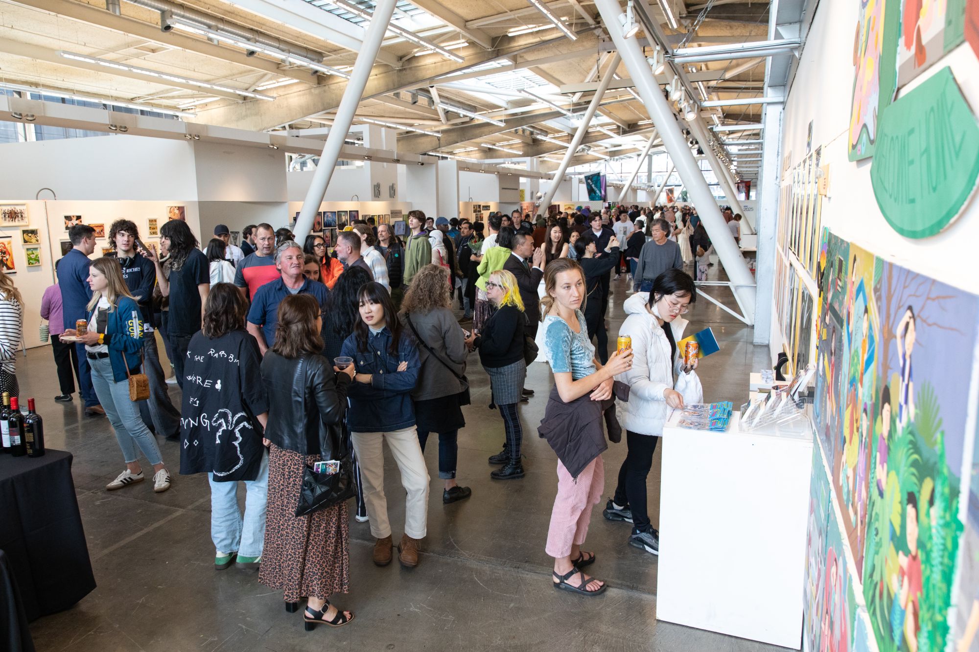 A large crowd of students, families, and guests fill the CCA Main Building gallery space during the BFA Illustration Senior Show opening, with colorful illustration work displayed on the walls.