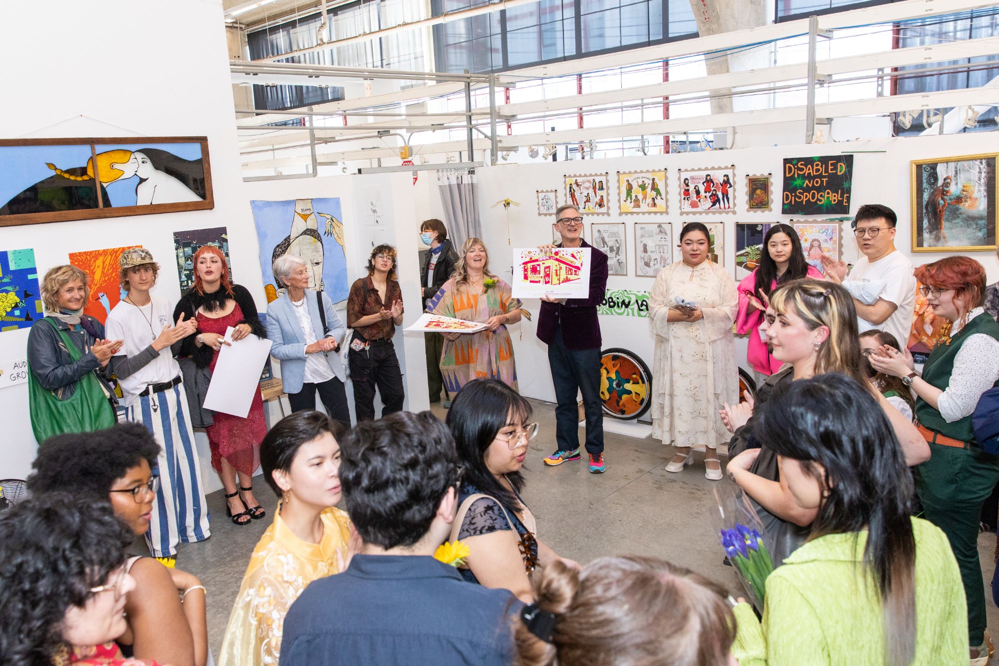 Students and faculty gather in a circle to celebrate during the BFA Illustration Senior Show, with an instructor holding a colorful printed illustration at the center.